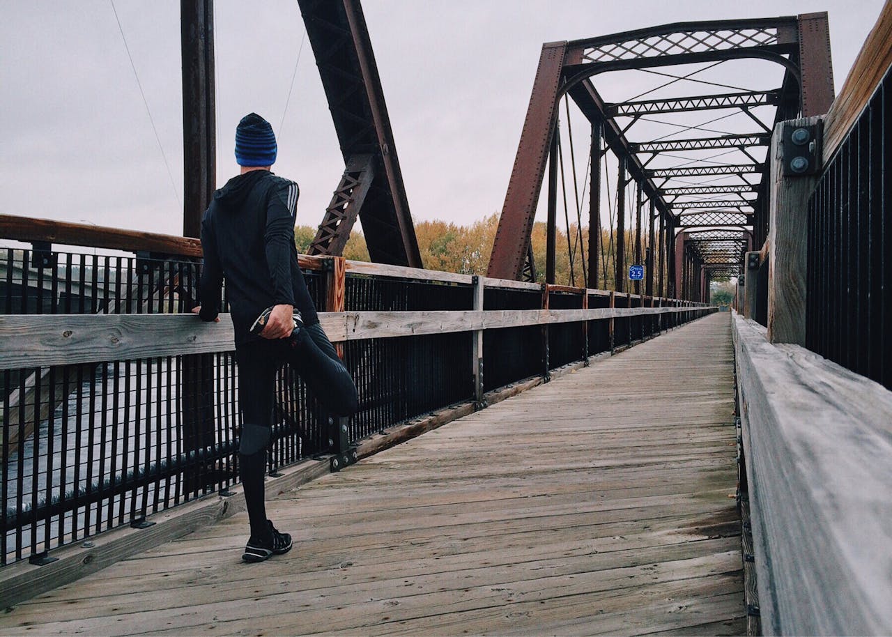 gallery-03 Man stretching on a wooden footbridge during a daytime run on an iron bridge, emphasizing fitness and recreation.
